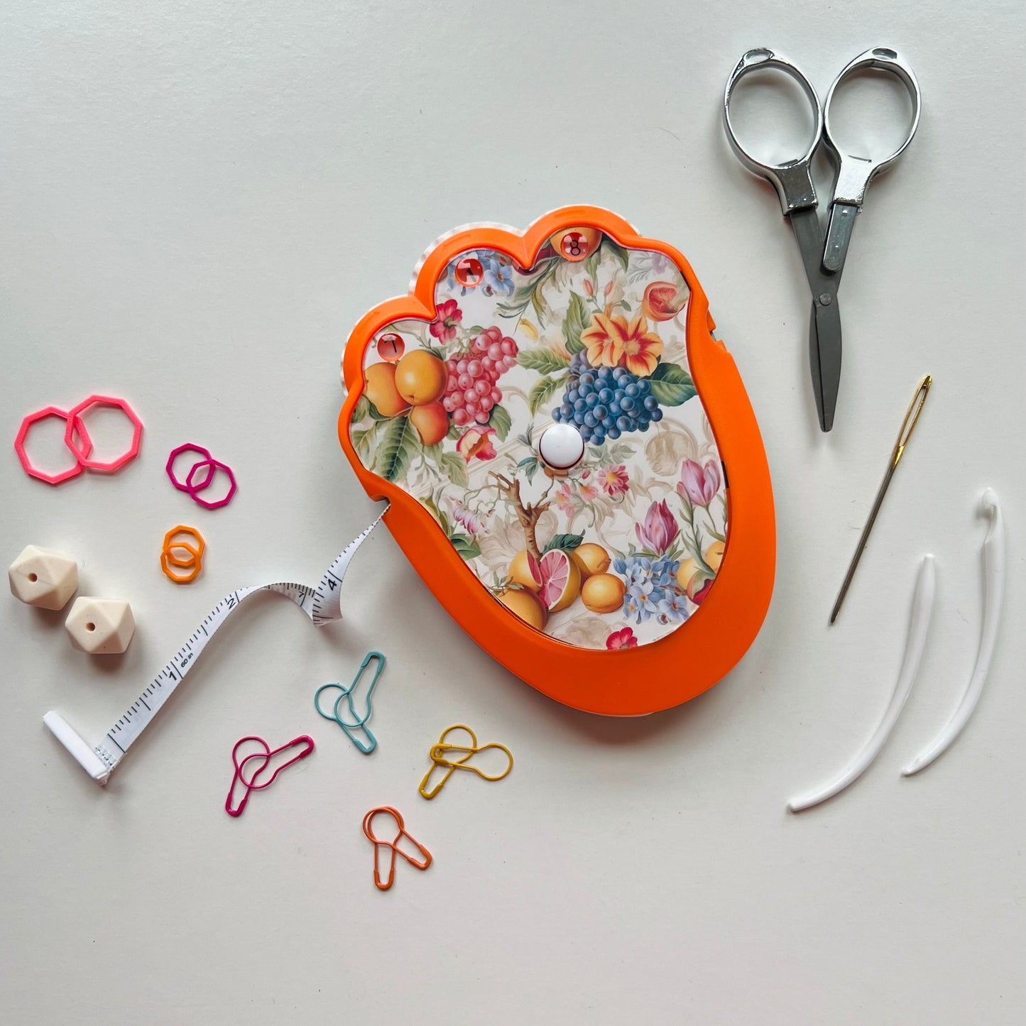 Decorative knitting kit with floral-patterned case, scissors, measuring tape, and various knitting tools on a light gray background.