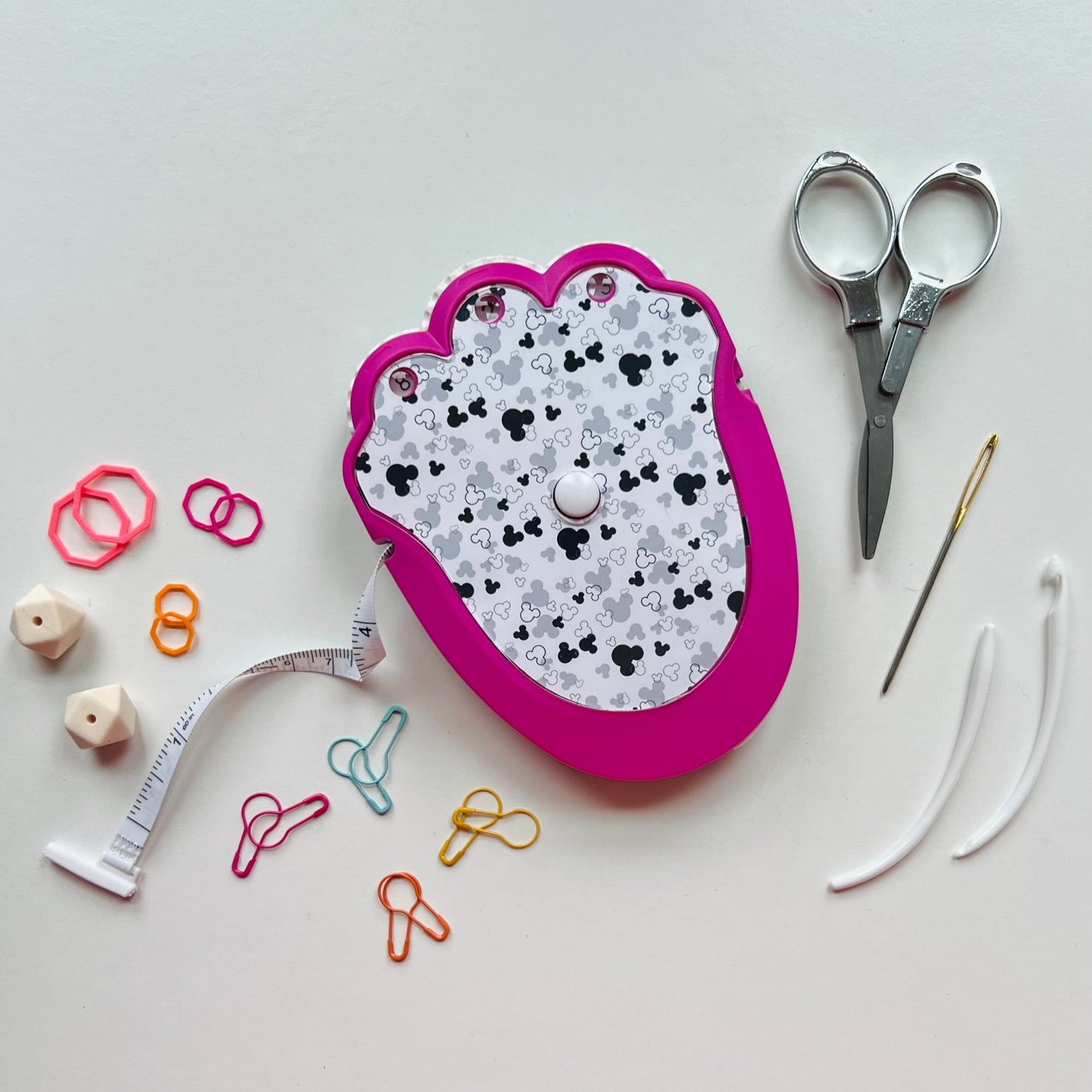Pink knitting kit with patterned lid, scissors, needle, thread, measuring tape, and colorful pins on a white background.