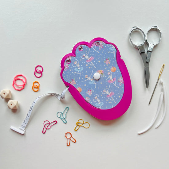 Pink knitting kit with scissors, measuring tape, and other tools on a light gray background