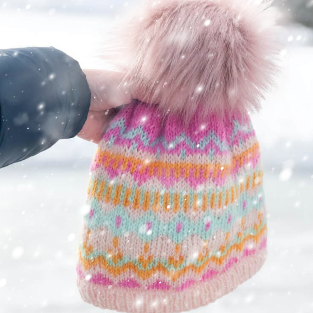 Colorful knitted beanie with a fluffy pom-pom held by a hand against a snowy background.
