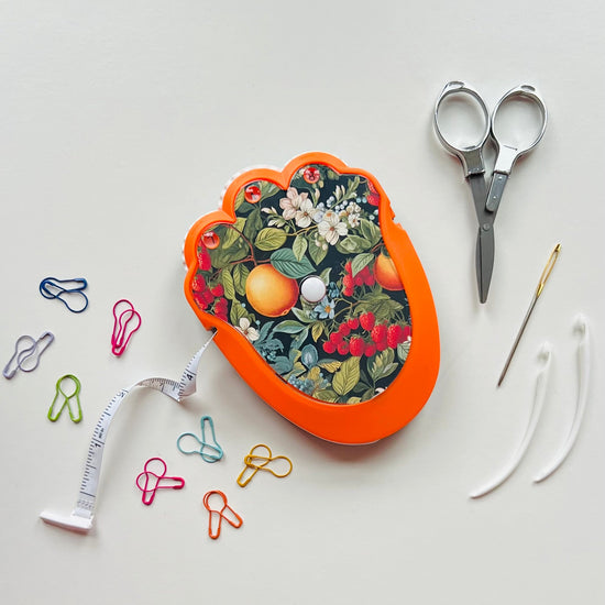 Decorative crocheting kit with floral design on an orange case, scissors, thread, and paper clips on a light gray background.